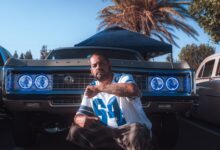 A man poses confidently in front of a classic car at an outdoor auto show, showcasing vibrant blue headlights.