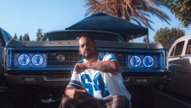 A man poses confidently in front of a classic car at an outdoor auto show, showcasing vibrant blue headlights.
