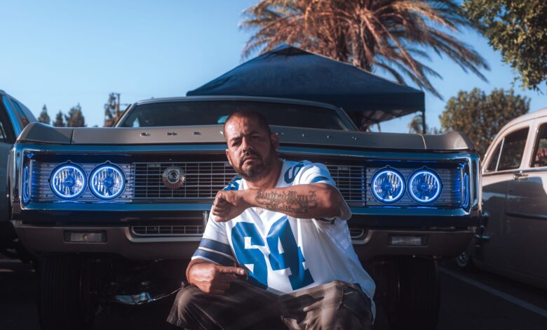 A man poses confidently in front of a classic car at an outdoor auto show, showcasing vibrant blue headlights.