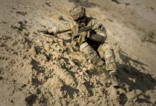 A soldier in full camouflage gear slides down a sandy slope during military training.