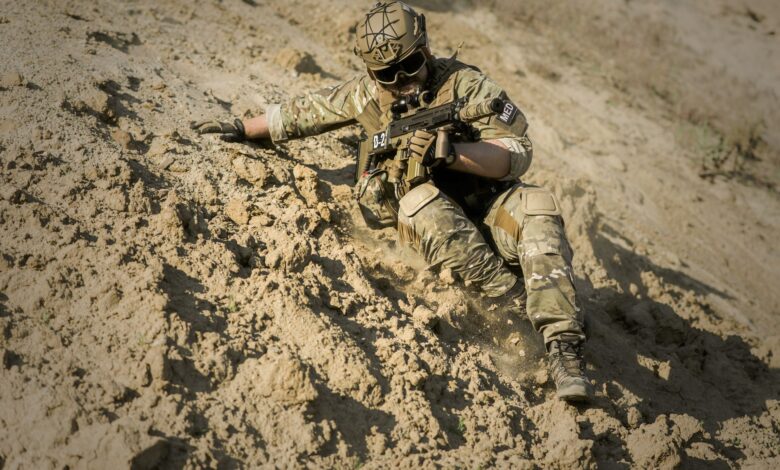 A soldier in full camouflage gear slides down a sandy slope during military training.