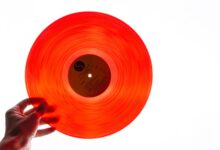 A bright orange vinyl record being held by a hand against a plain white background, showcasing its translucency.