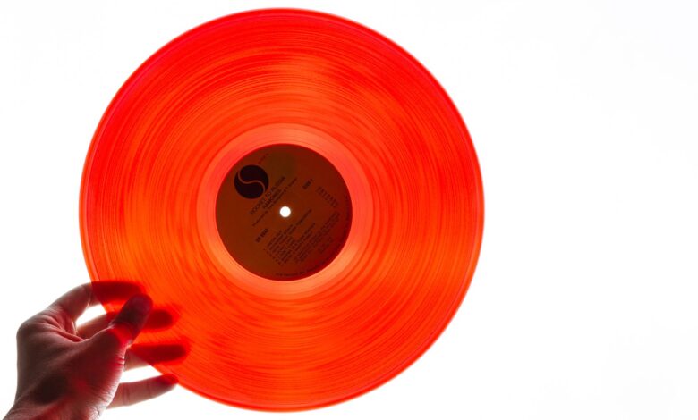 A bright orange vinyl record being held by a hand against a plain white background, showcasing its translucency.