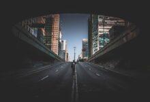A solitary figure walks on a city street beneath tall skyscrapers on a bright day.