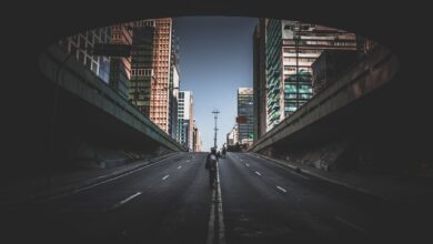 A solitary figure walks on a city street beneath tall skyscrapers on a bright day.
