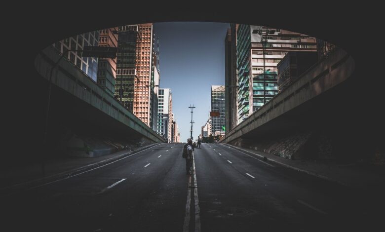 A solitary figure walks on a city street beneath tall skyscrapers on a bright day.