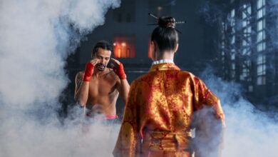 Two martial artists in traditional attire face off in a dramatic, smoke-filled arena.