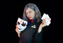 Side view of serious young female casino dealer in classy suit with bow tie holding face mask and cards in hands and looking at camera against black background