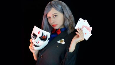 Side view of serious young female casino dealer in classy suit with bow tie holding face mask and cards in hands and looking at camera against black background