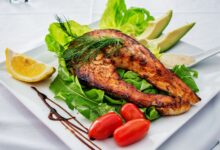 Delicious grilled salmon steak served with avocado, lettuce, cherry tomatoes, and lemon slice on a white plate.