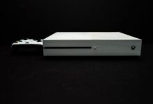 Close-up of a white gaming console with a black controller on a dark background.