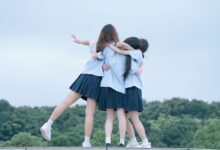 Three teenage girls in school uniforms hugging outdoors on a cloudy day.