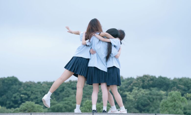 Three teenage girls in school uniforms hugging outdoors on a cloudy day.
