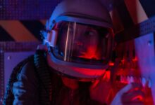 A woman astronaut wearing a spacesuit in a control room lit by red lights, looks focused.