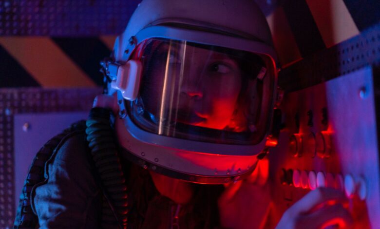 A woman astronaut wearing a spacesuit in a control room lit by red lights, looks focused.