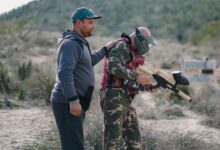 Two men playing paintball outdoors, one wearing camouflage and a mask, in a rural setting.