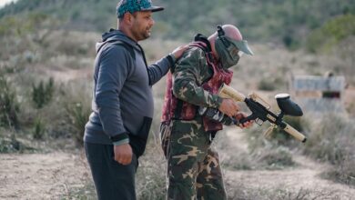 Two men playing paintball outdoors, one wearing camouflage and a mask, in a rural setting.