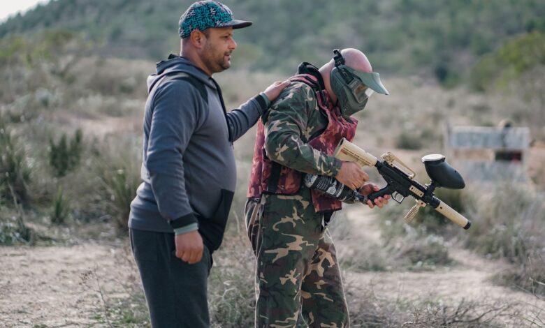 Two men playing paintball outdoors, one wearing camouflage and a mask, in a rural setting.