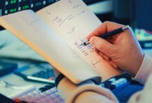 A hand writing in a notebook during stock market analysis on a desk.