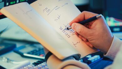 A hand writing in a notebook during stock market analysis on a desk.