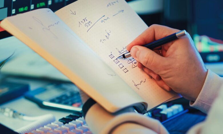 A hand writing in a notebook during stock market analysis on a desk.