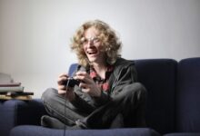 Enthusiastic teen boy in casual wear and eyeglasses sitting on blue sofa with legs crossed and playing video game with joystick at home