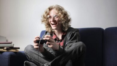 Enthusiastic teen boy in casual wear and eyeglasses sitting on blue sofa with legs crossed and playing video game with joystick at home