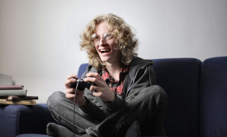 Enthusiastic teen boy in casual wear and eyeglasses sitting on blue sofa with legs crossed and playing video game with joystick at home