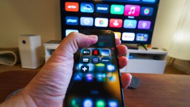 Close-up of a hand holding a phone displaying streaming apps in front of a TV with multiple app icons.