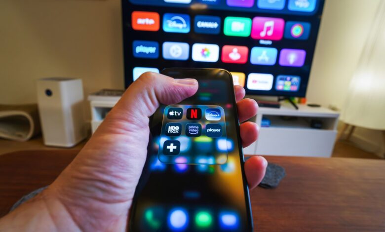 Close-up of a hand holding a phone displaying streaming apps in front of a TV with multiple app icons.