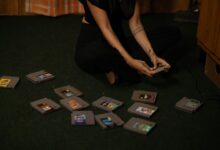Adult woman sitting cross-legged, holding a vintage game controller, surrounded by retro game cartridges.