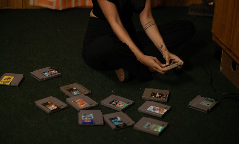 Adult woman sitting cross-legged, holding a vintage game controller, surrounded by retro game cartridges.