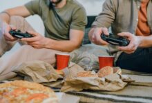Two friends playing video games and enjoying burgers and pizza indoors with red cups.