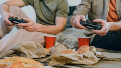 Two friends playing video games and enjoying burgers and pizza indoors with red cups.