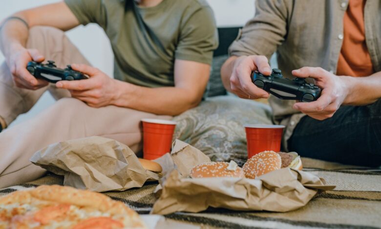 Two friends playing video games and enjoying burgers and pizza indoors with red cups.