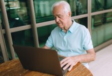 Elderly man sitting at a wooden table, working on a laptop indoors.
