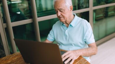 Elderly man sitting at a wooden table, working on a laptop indoors.