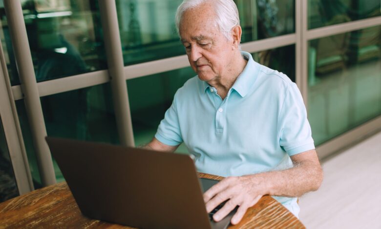 Elderly man sitting at a wooden table, working on a laptop indoors.