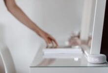 A smart speaker with a digital clock display placed on a minimalist desk setting, hand in background.