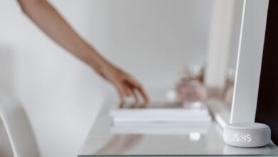 A smart speaker with a digital clock display placed on a minimalist desk setting, hand in background.