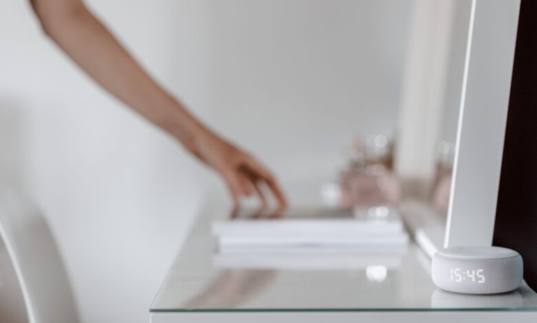A smart speaker with a digital clock display placed on a minimalist desk setting, hand in background.