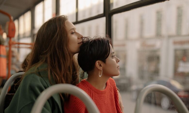 Two young women share an intimate moment on a bus, gazing thoughtfully outside.