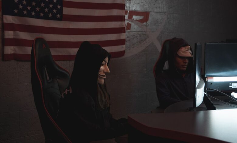 Two masked individuals in a dark room working on computers, symbolizing cyber security threats.