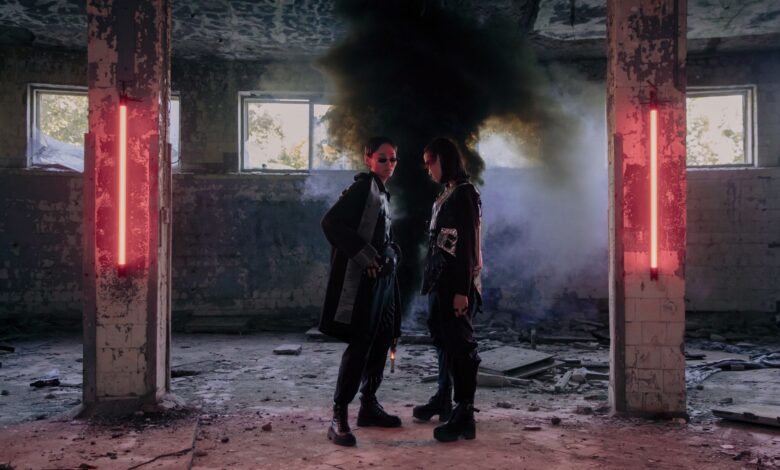 Two models in a dystopian setting with neon lights and smoke, showcasing futuristic fashion.