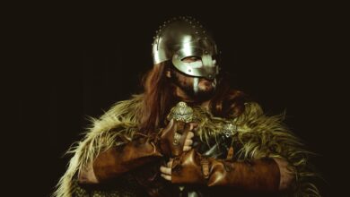A viking warrior wearing a helmet and armor with fur covering against a dark background.