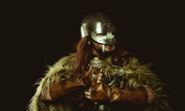 A viking warrior wearing a helmet and armor with fur covering against a dark background.