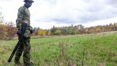 A soldier in camouflage with a rifle standing on a grassy field under a cloudy sky.