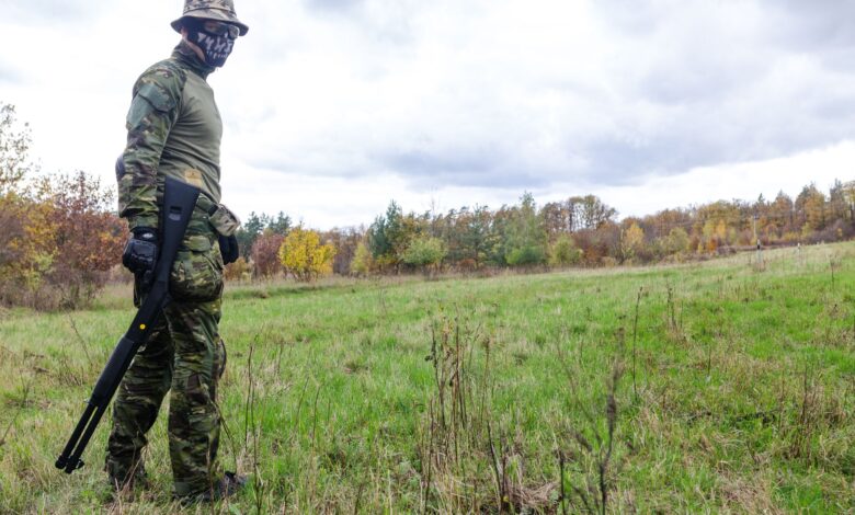 A soldier in camouflage with a rifle standing on a grassy field under a cloudy sky.