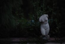 A broken stone torso statue surrounded by dark foliage, evoking mystery.
