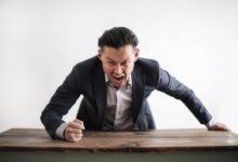 Expressive angry businessman in formal suit looking at camera and screaming with madness while hitting desk with fist